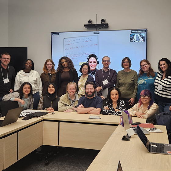 A group of workshop participants poses together in a conference room, with laptops on the tables and a large screen displaying a virtual attendee in the background.