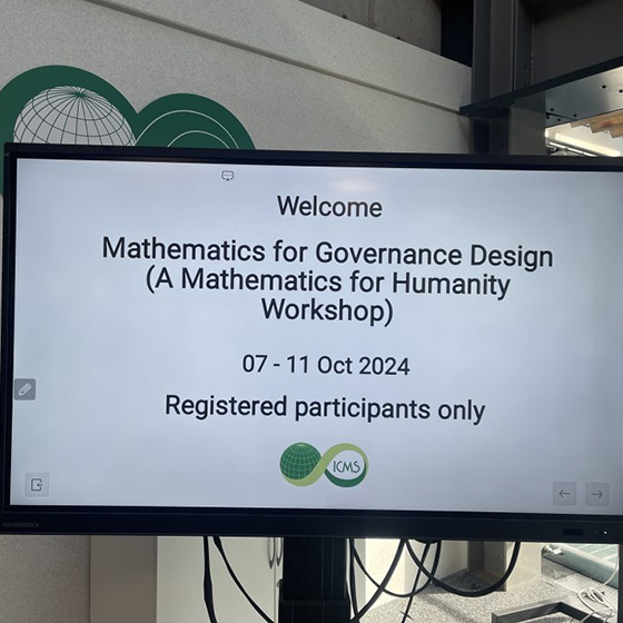 A monitor displays a welcome screen for the Mathematics for Governance Design workshop, a Mathematics for Humanity event held October 7–11, 2024, for registered participants only, with the ICMS logo shown at the bottom.