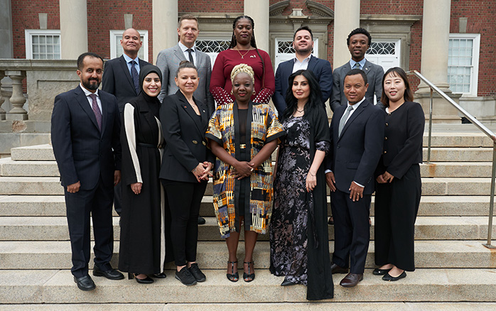 A group of eleven people are standing on the steps of a brick building with large windows and white columns. They are all dressed in formal attire, and the setting suggests a professional or ceremonial occasion.