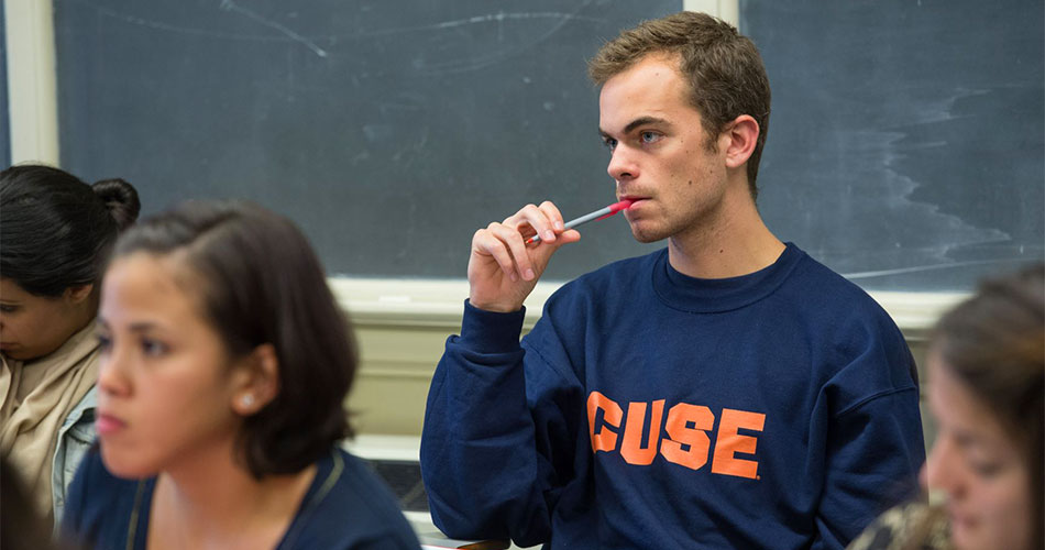 Male student in a classroom with a cuse sweatshirt and pen in hand.