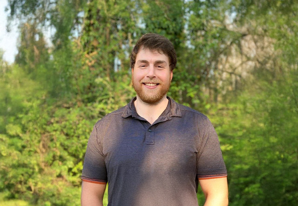 A person standing outdoors in a lush, green area with trees and bushes in the background. They are smiling and wearing a short-sleeved shirt.