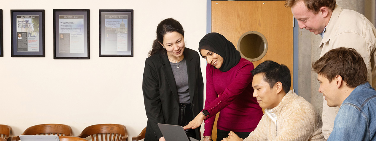 A group of people are gathered around a table looking at a laptop. There are framed certificates on the wall behind them. The setting appears to be a casual meeting room.