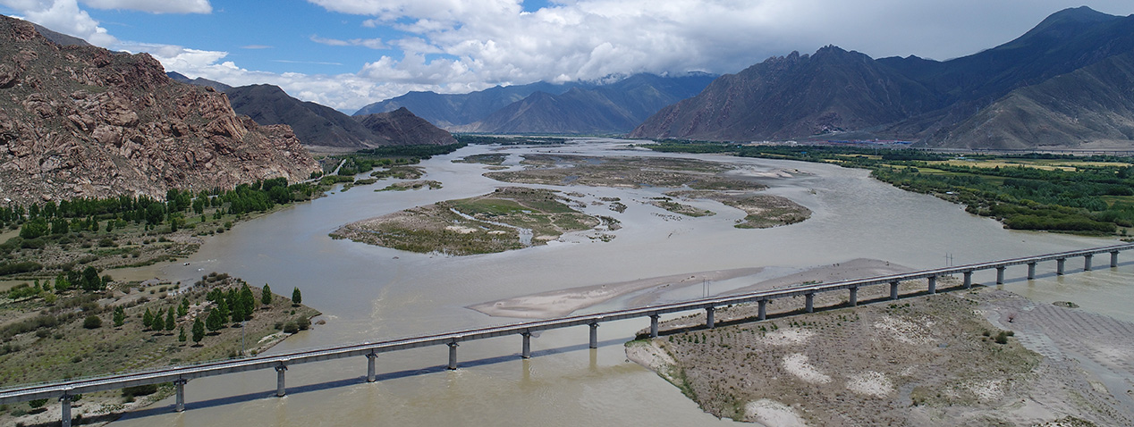 A wide river flows under a long bridge, surrounded by a landscape of mountains and scattered greenery. The scene is expansive with partly cloudy skies above.