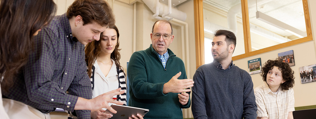 Professor speaking with hands raised while students gather around a tablet.