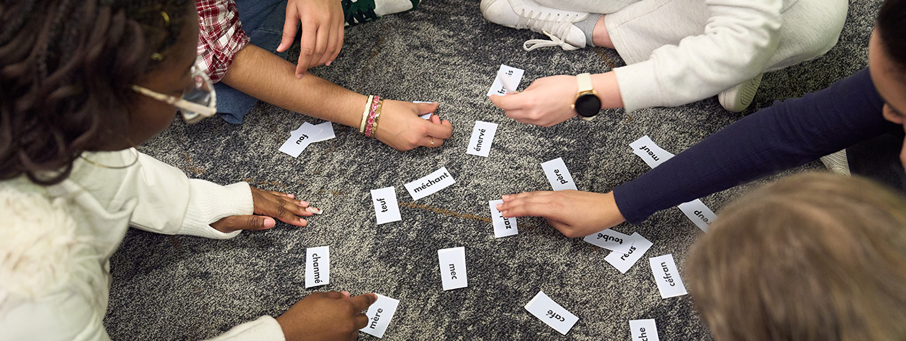 Individuals are seated on the floor, collaboratively arranging word cards on a dark carpet. Hands are visible reaching in, with some wearing casual clothing and accessories like bracelets and a smartwatch.
