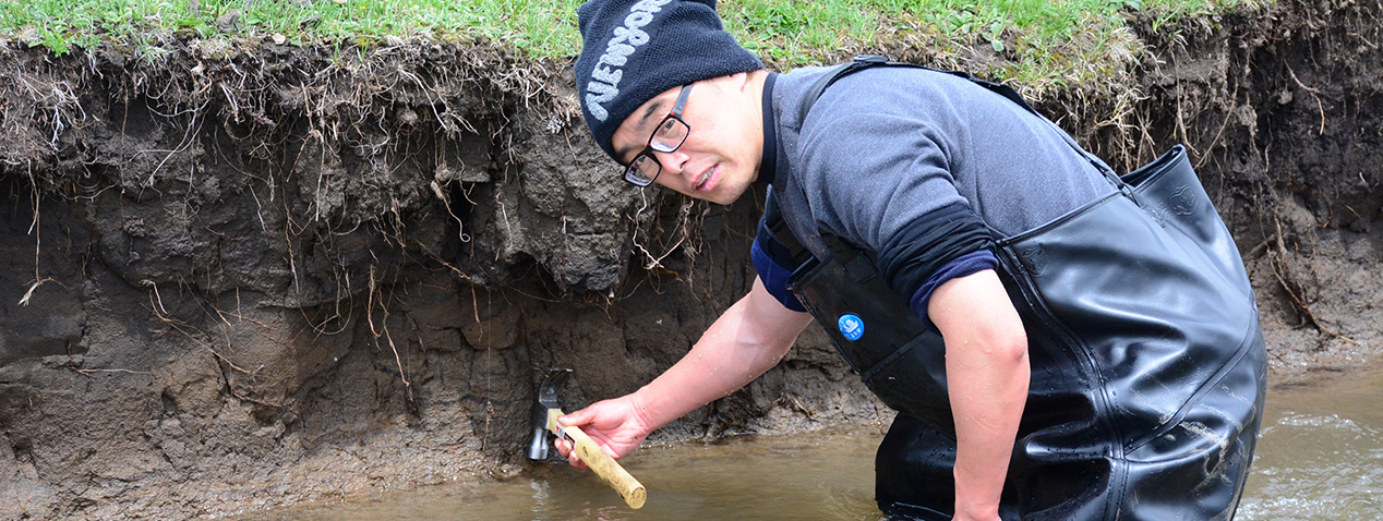 A person wearing a beanie and waders is examining a riverbank with a hammer. They are standing in shallow water. The grassy bank shows exposed soil layers.