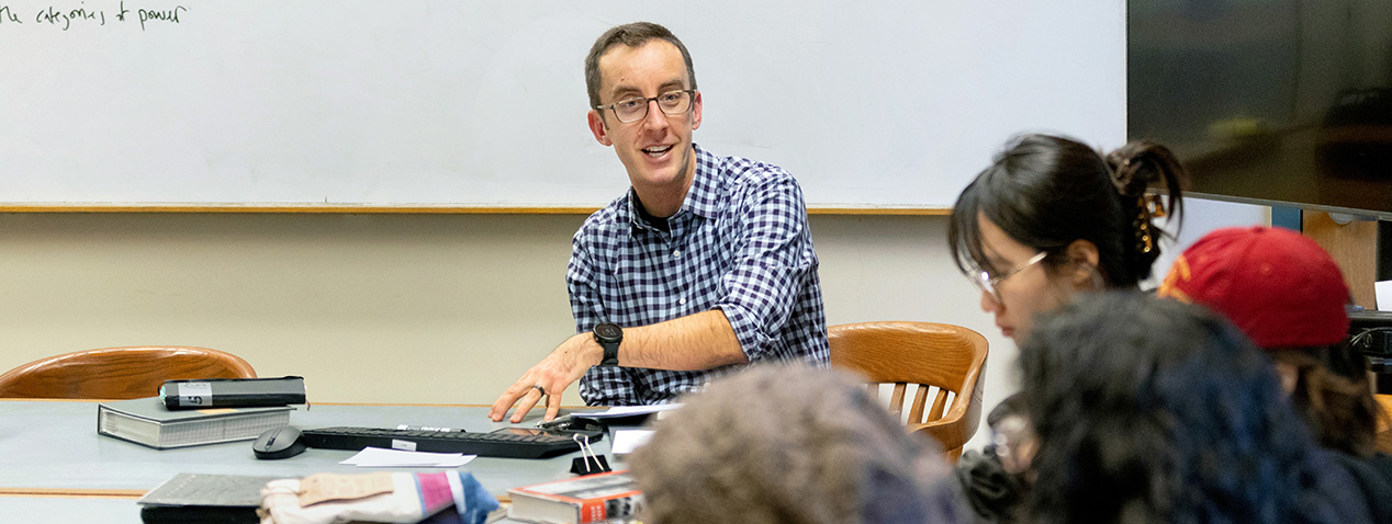 A person wearing glasses sits at a table with open books and engages with a group of individuals in a classroom setting. A large whiteboard is visible in the background.