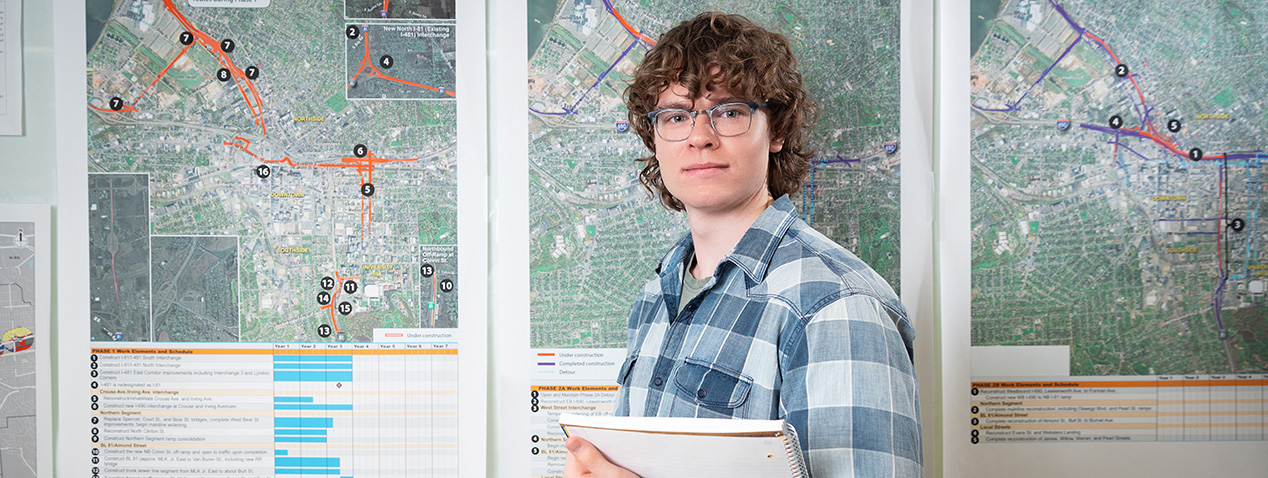 A person holding a clipboard stands in front of detailed city maps and charts. The maps display various routes and geographic information. The person is wearing glasses and a plaid shirt.