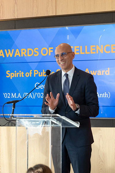 A person in a suit stands at a podium, smiling and gesturing, with a microphone in front of a screen displaying