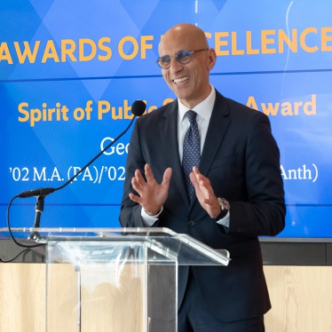 A person in a suit stands at a podium, smiling and gesturing, with a microphone in front of a screen displaying "Awards of Excellence" and "Spirit of Public Service Award.