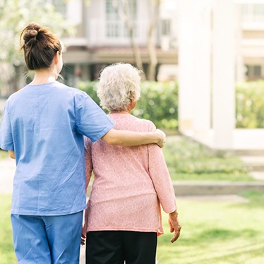 Caregiver in scrubs walks arm-in-arm with an older adult through a sunny courtyard garden.