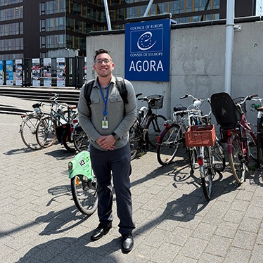 Person smiling in front of the Council of Europe Agora building, with bicycles parked nearby.