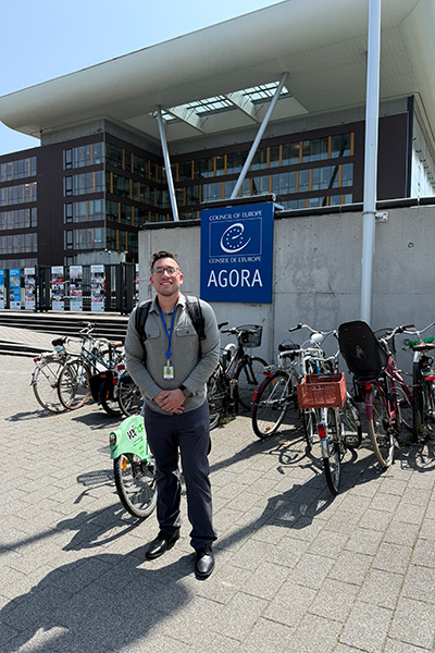 Person smiling in front of the Council of Europe Agora building, with bicycles parked nearby.
