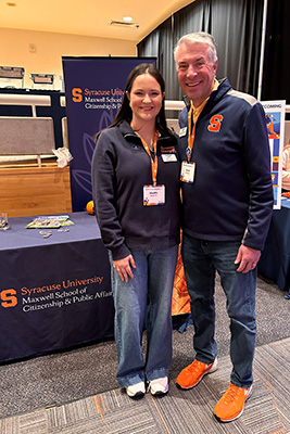 Two individuals posing at a Syracuse University event, standing in front of a Maxwell School of Citizenship & Public Affairs banner. Both are smiling and wearing Syracuse University apparel.