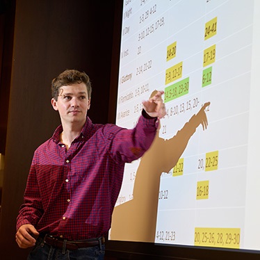 A person stands in front of a presentation screen, pointing at highlighted data on a chart. The chart displays numbers and text, some of which are highlighted. The setting appears to be a classroom or lecture hall.