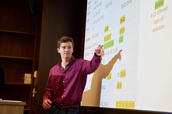 A person stands in front of a presentation screen, pointing at highlighted data on a chart. The chart displays numbers and text, some of which are highlighted. The setting appears to be a classroom or lecture hall.