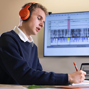 A person wearing orange headphones and a navy sweater sits at a desk, writing on a tablet with a stylus. A large screen in the background displays a data visualization with waveform and spectrogram-like graphics.