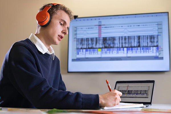 A person wearing orange headphones and a navy sweater sits at a desk, writing on a tablet with a stylus. A large screen in the background displays a data visualization with waveform and spectrogram-like graphics.