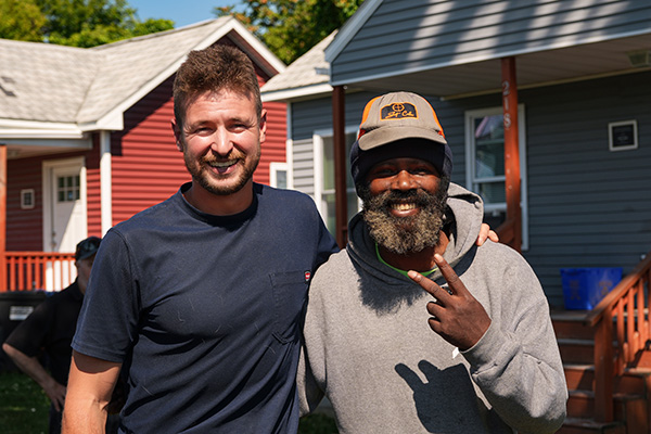 Two people smile together outside small homes, one giving a peace sign.