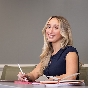 A professional sits at a table in an office setting, holding a pen and smiling while looking away from a notebook and a laptop.