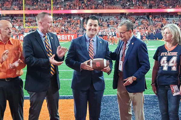 Five individuals are standing on a football field: one is being presented with an award by another, while three others applaud. They are dressed in business casual attire with some wearing Syracuse University logos.
