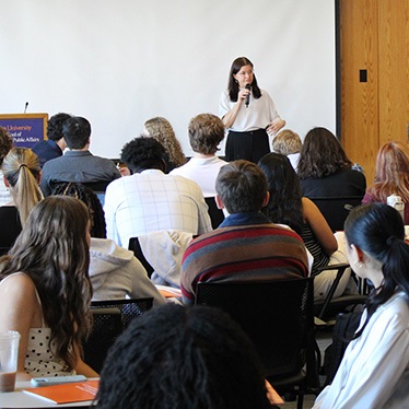 A woman stands at the front of a classroom speaking into a microphone while addressing an audience of seated attendees.
