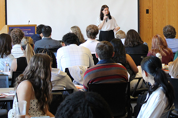 A woman stands at the front of a classroom speaking into a microphone while addressing an audience of seated attendees.