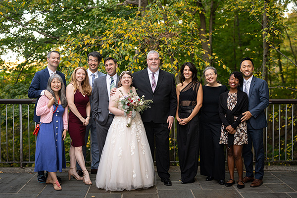 Group of ten people in formal attire, including a bride holding a bouquet, posing for a photo outdoors with trees in autumn colors in the background.