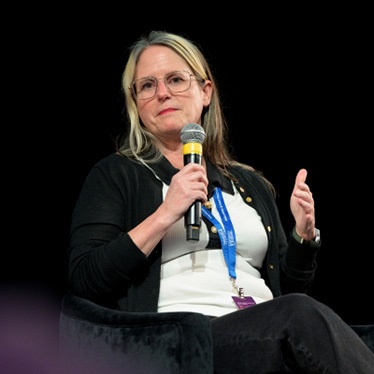 Person speaking into a microphone at a conference, seated onstage.