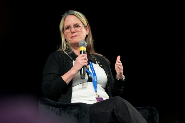Person speaking into a microphone at a conference, seated onstage.