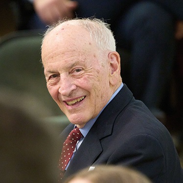 An person in a dark suit and red tie smiles warmly, photographed in what appears to be a lecture hall or auditorium setting.