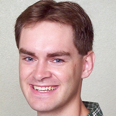 Smiling person with short brown hair and a green plaid collar in front of a neutral backdrop.