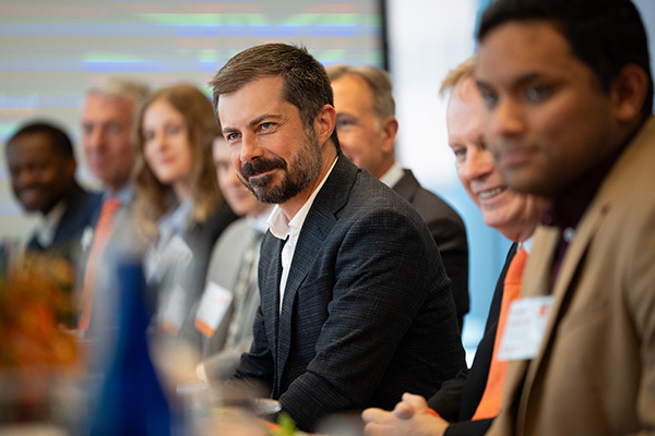 Group of professionals, including former US Transportation Secretary Pete Buttigieg, sitting at a conference table, engaged in a discussion.