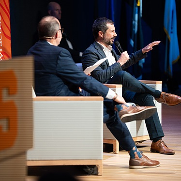 Two individuals engaged in conversation onstage at an event, with one person gesturing while seated.