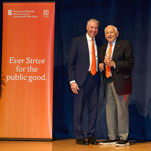 Two individuals standing on stage at a Syracuse University event, smiling and dressed in formal attire, beside a banner with the text