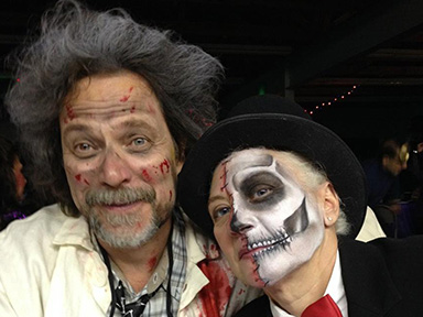 Two people at a costume party, one with wild hair and fake blood on their face, the other wearing skeleton makeup and a top hat.