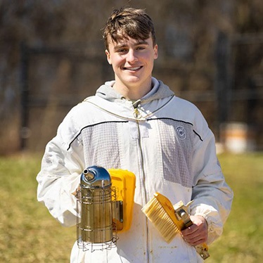 A person stands outdoors holding beekeeping equipment, smiling and wearing a light jacket with a visible logo.