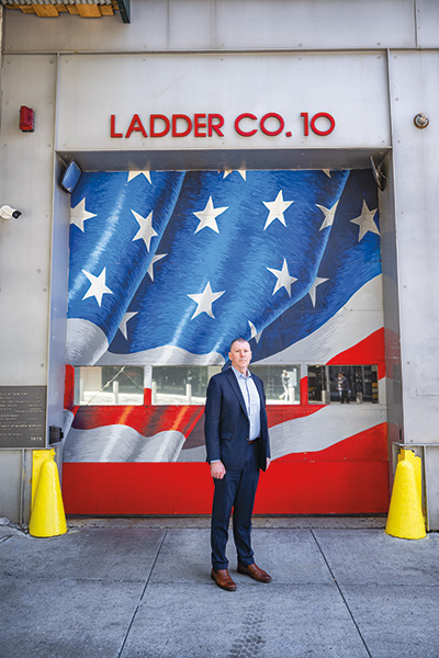 Person in a business suit standing in front of Ladder Co. 10 fire station, which features a large American flag mural.