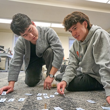 Two individuals are kneeling on a carpeted floor, engaged in sorting or arranging small cards. They appear focused on their task. The setting seems to be an indoor office or classroom environment with desks and chairs in the background.