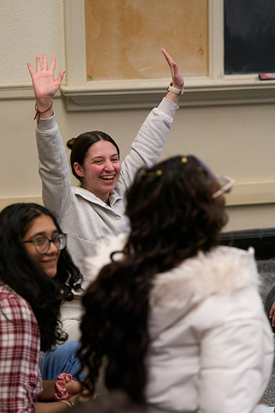 A person with raised hands smiles and engages in a classroom setting with two others. There is a chalkboard and a bulletin board in the background.