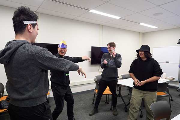 A group of people in a room having fun. They are engaged in an activity with two wearing playful paper crowns. The room has white walls, two flat screen TVs, and a whiteboard. The atmosphere is casual and lively.