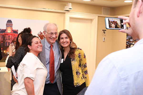 A group of people smiling for a photo, with one person taking the picture on a smartphone. They are indoors, and in the background, there is a picture of a building.