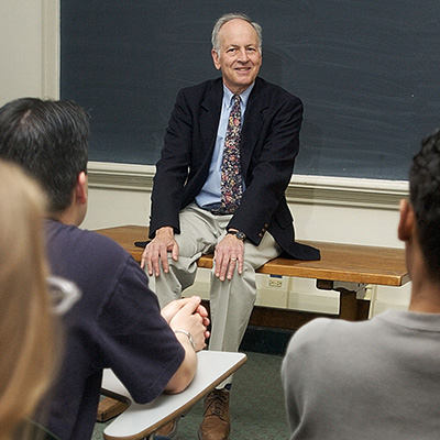 A person sits on a desk at the front of a classroom, smiling and engaged in conversation with students seated in front of him. A chalkboard is visible in the background.