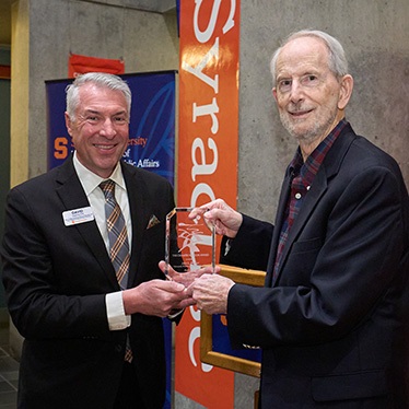 Two individuals exchanging an award at a Syracuse University event, with banners and university imagery in the background.