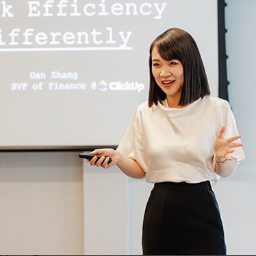 A person presents in front of a projection screen displaying the slide title "Think Efficiency Differently" during a speaking engagement.