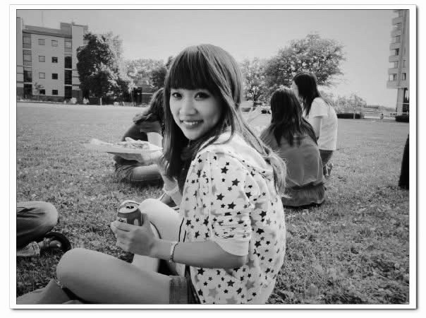 A black-and-white photo of a person sitting on a campus lawn, smiling over their shoulder at the camera while holding a can and a plate of food, with other students and university buildings visible in the background.
