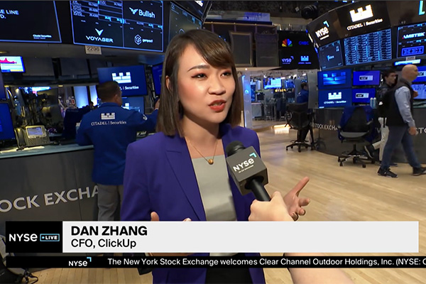 A person speaks into a NYSE microphone on the floor of the New York Stock Exchange, surrounded by trading screens and financial displays.
