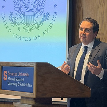 An individual speaking at a podium labeled 'Syracuse University Maxwell School of Citizenship & Public Affairs' with the United States seal projected in the background.