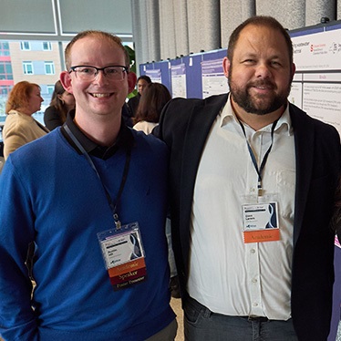 Two people stand together and smile for a photo at an academic conference, with research poster displays and attendees visible in the background. Both wear conference lanyards; one wears a blue sweater and glasses, the other a dark blazer.