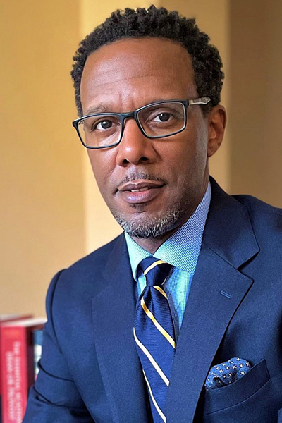 A person in a navy suit and striped tie, glasses, bookshelf in background.